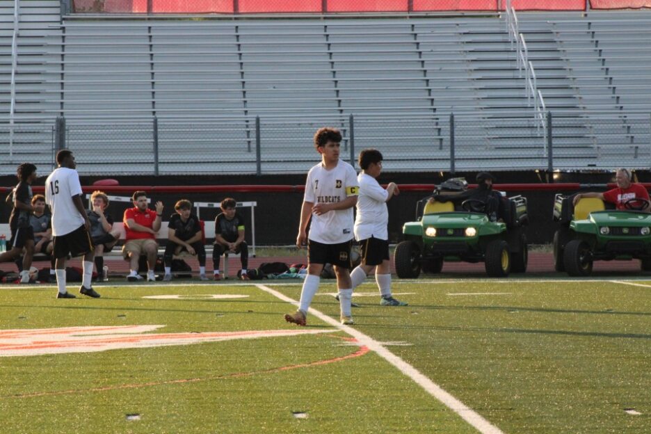 Two soccer players in white uniforms stand on a field, with empty stadium bleachers and team members seated in the background.