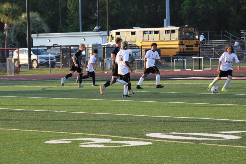 Soccer players in black and white uniforms run on a field during a game, with a yellow school bus and cars in the background.