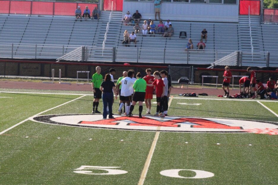 Soccer players and referees gathered at midfield on a sports field, with bleachers and a few spectators in the background.
