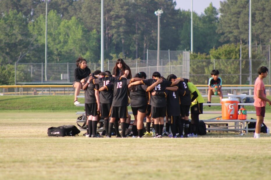 A soccer team in black uniforms huddles together on a field, with benches, bags, water coolers, and people sitting and standing nearby.