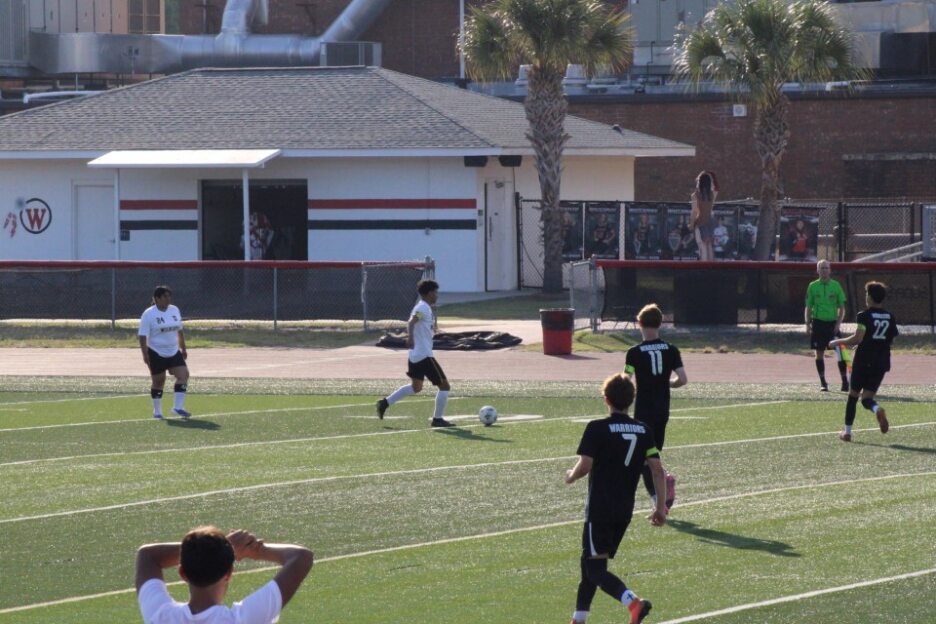 Soccer players in black and white uniforms play a match on a sunny outdoor field, with buildings and trees visible in the background.