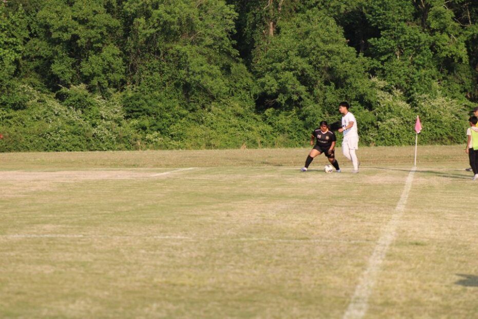 Two soccer players compete for the ball near the corner flag on a grassy field, with trees in the background.