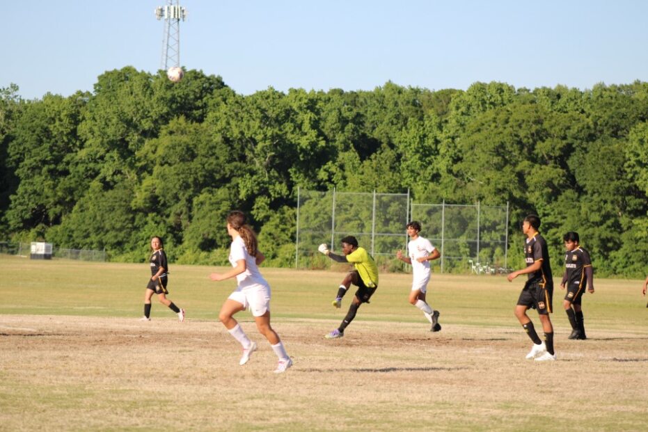 A soccer goalkeeper in yellow kicks the ball while players in white and black uniforms run on a grassy field with trees in the background.