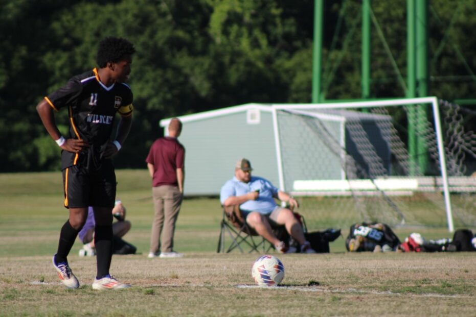 A soccer player in a black uniform stands on the field near a ball, with people sitting and a goalpost in the background.