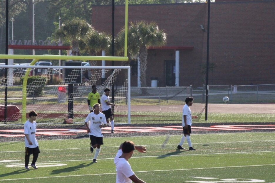 Soccer players on a field during a game, with a goalkeeper in front of the goal and a school building in the background.