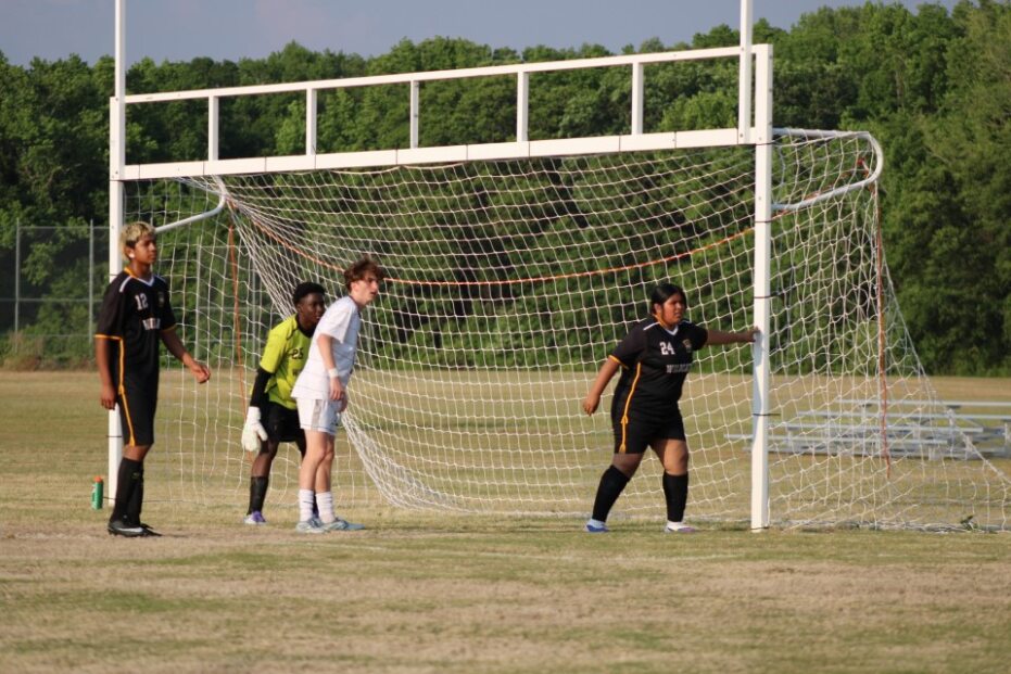 Four soccer players stand inside a goal, preparing for a defensive play during an outdoor match on a grassy field with trees in the background.