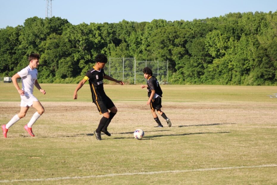 Three soccer players compete for the ball on a grassy field with a background of green trees under a clear blue sky.