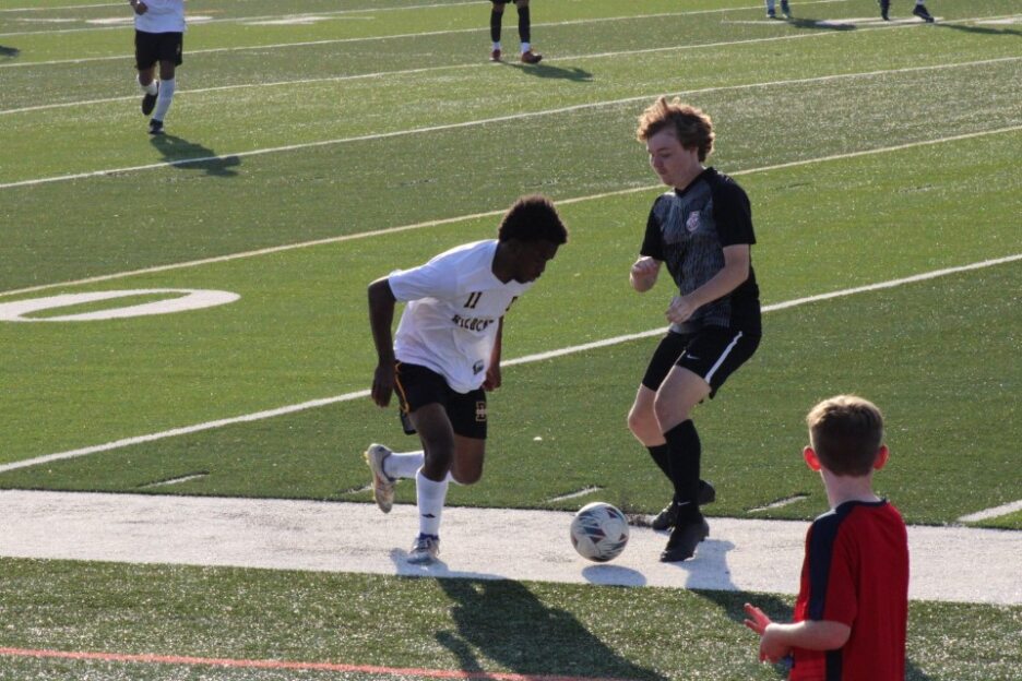 Two youth soccer players compete for the ball near the sideline on a sunny day, with other players and a referee visible in the background.