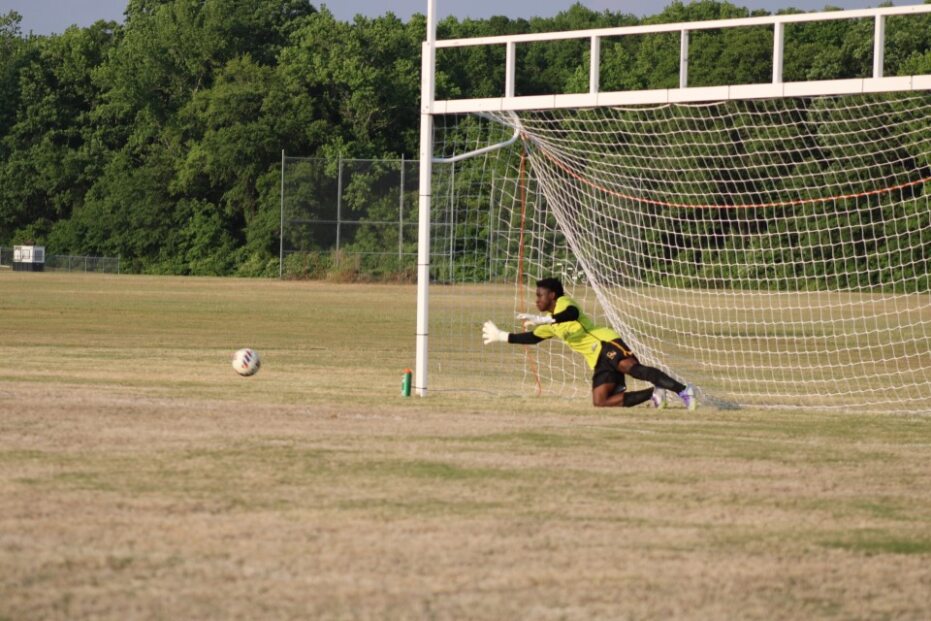 A soccer goalkeeper in yellow dives but misses the ball, which enters the goal on a grassy outdoor field with trees in the background.
