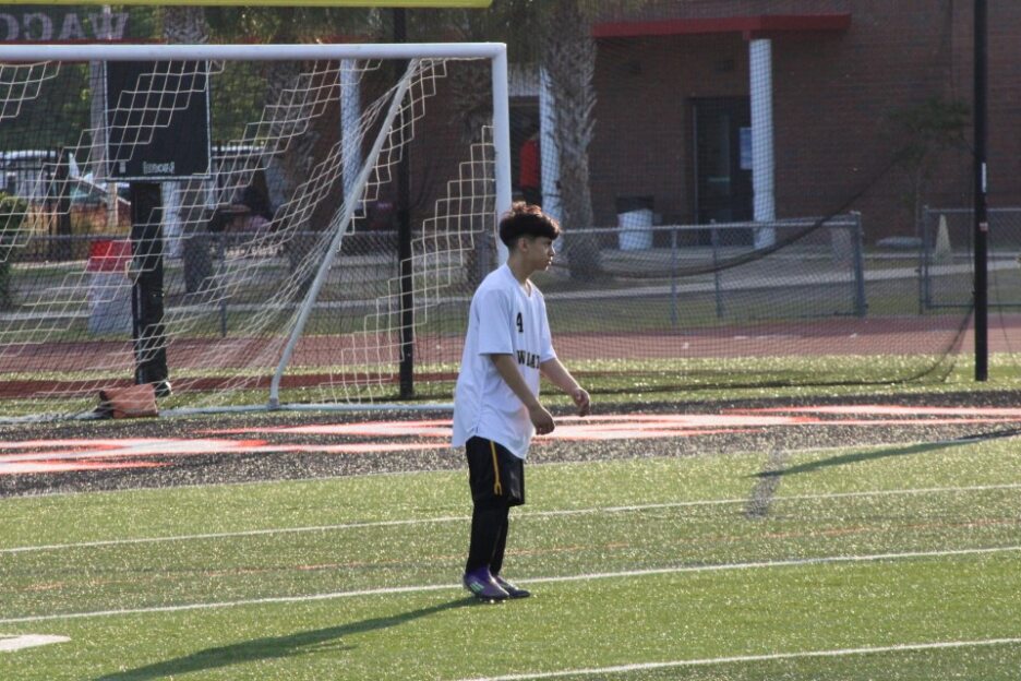 A boy in a white shirt and black pants stands in front of a soccer goal on a grassy field.