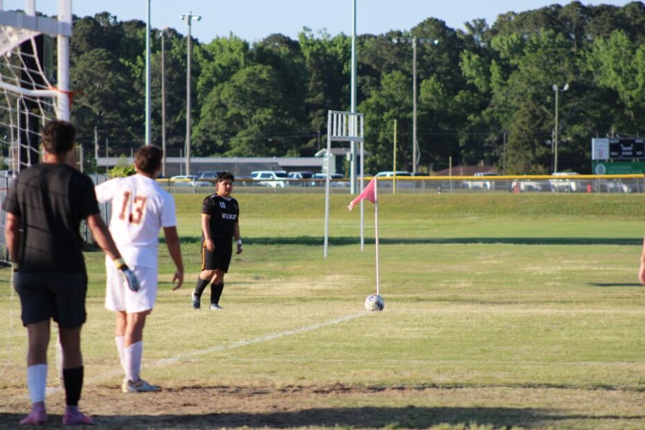 A soccer player prepares to take a corner kick during a game, with defenders and a goalkeeper waiting near the goal.