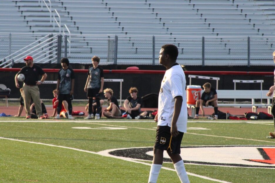 A soccer player in a white jersey stands on a field, with teammates and empty bleachers in the background.