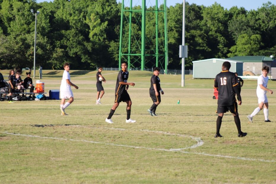 Soccer players in black and white uniforms on a grassy field, with trees, a green structure, and benches visible in the background under daylight.