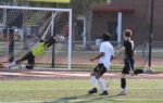 A soccer goalie dives to block a ball heading towards the goal, while two players watch the action on a grassy field.