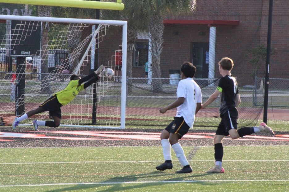 A soccer goalie dives to block a ball heading towards the goal, while two players watch the action on a grassy field.