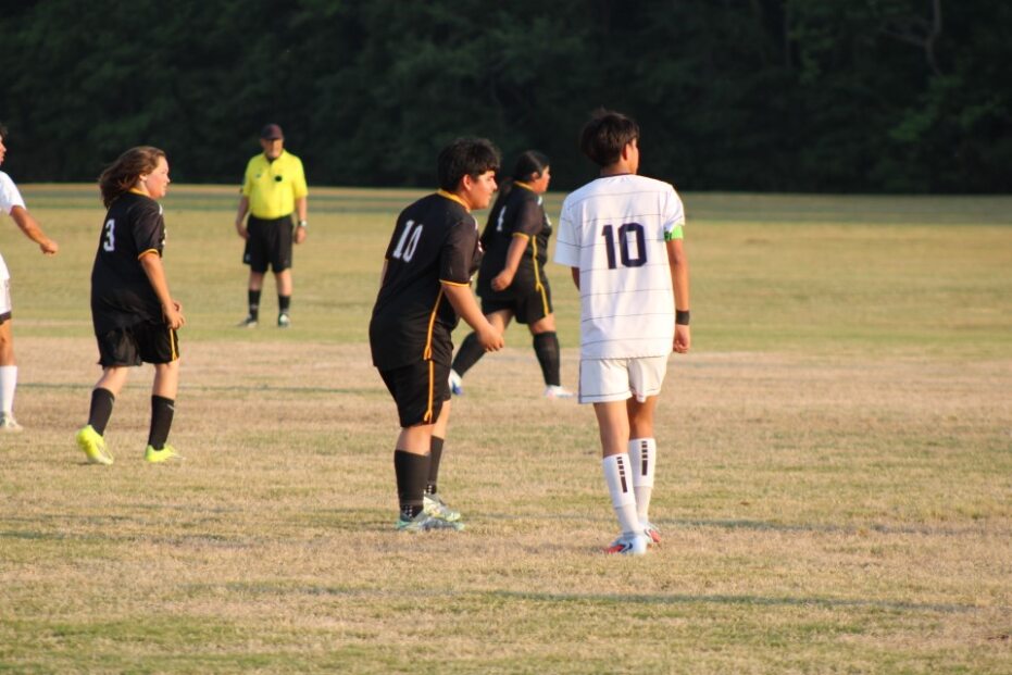 Soccer players on a field, with one team in black and another in white, and a referee in the background.