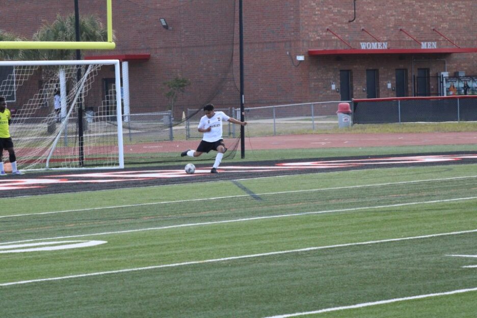 A soccer player in a white jersey kicks a ball near a goal on an outdoor field with a school building in the background.