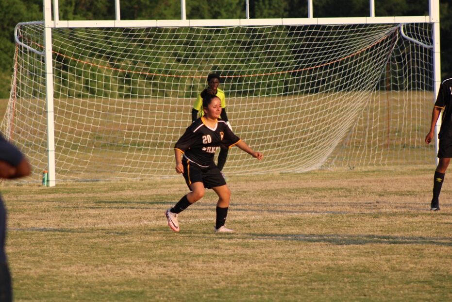 Soccer player in black uniform running on field with goal and net in background, two other players visible.