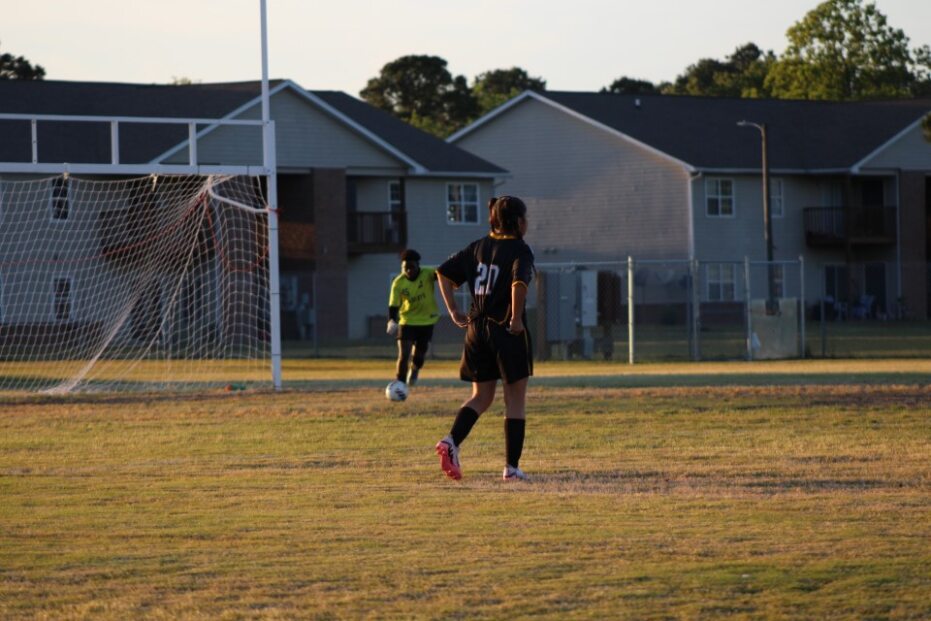 A soccer goalie preparing to kick the ball while another player in a black uniform stands near the goalpost on a grassy field.