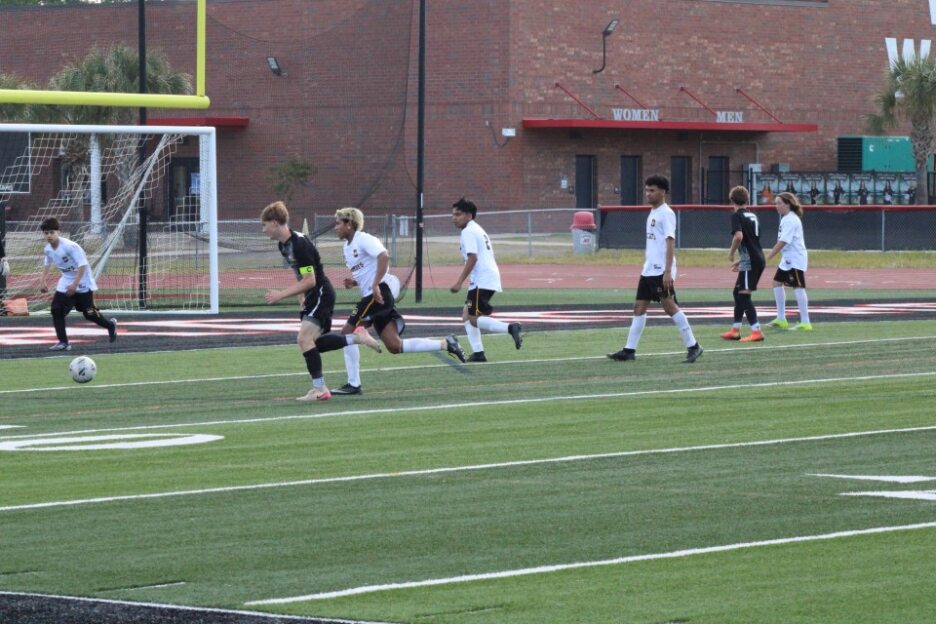 Soccer players in black and white uniforms running on a field near a goalpost, with a brick building in the background.