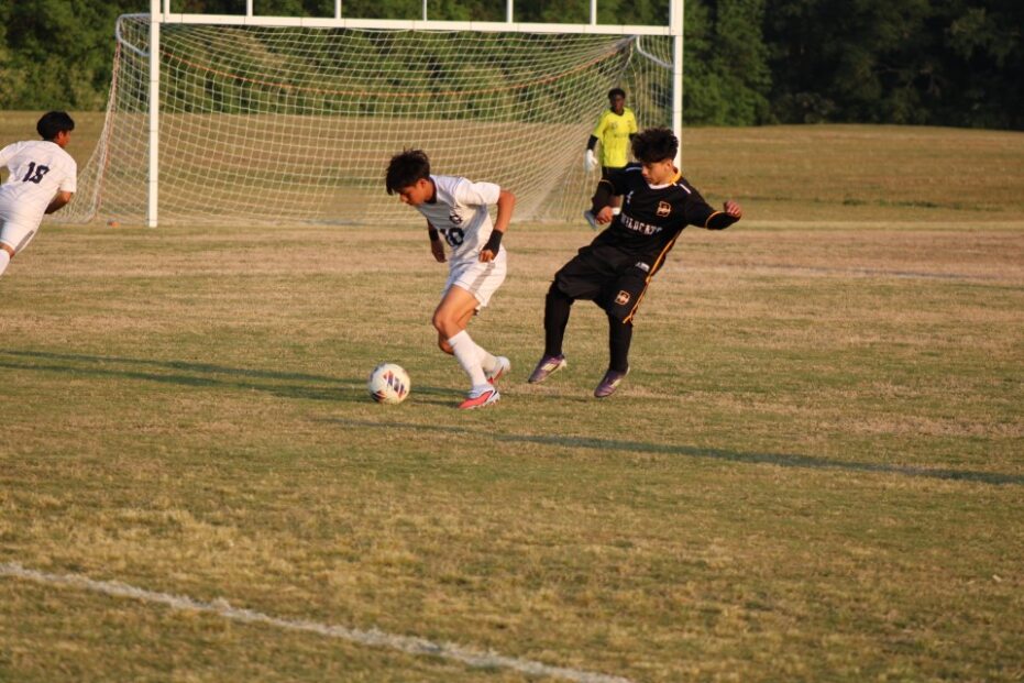 Two youth soccer players chase the ball near the goal, with a goalkeeper and another player in the background on a grassy field.