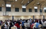 Large group of people seated at round tables in a gymnasium, participating in an indoor event or gathering under high ceilings with visible basketball hoops.