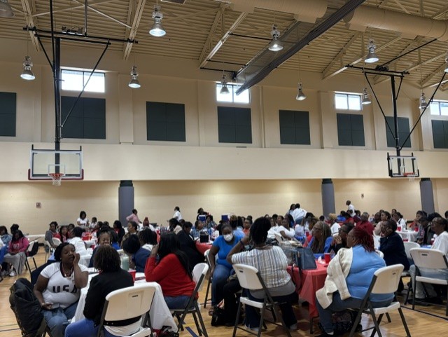 Large group of people seated at round tables in a gymnasium, participating in an indoor event or gathering under high ceilings with visible basketball hoops.