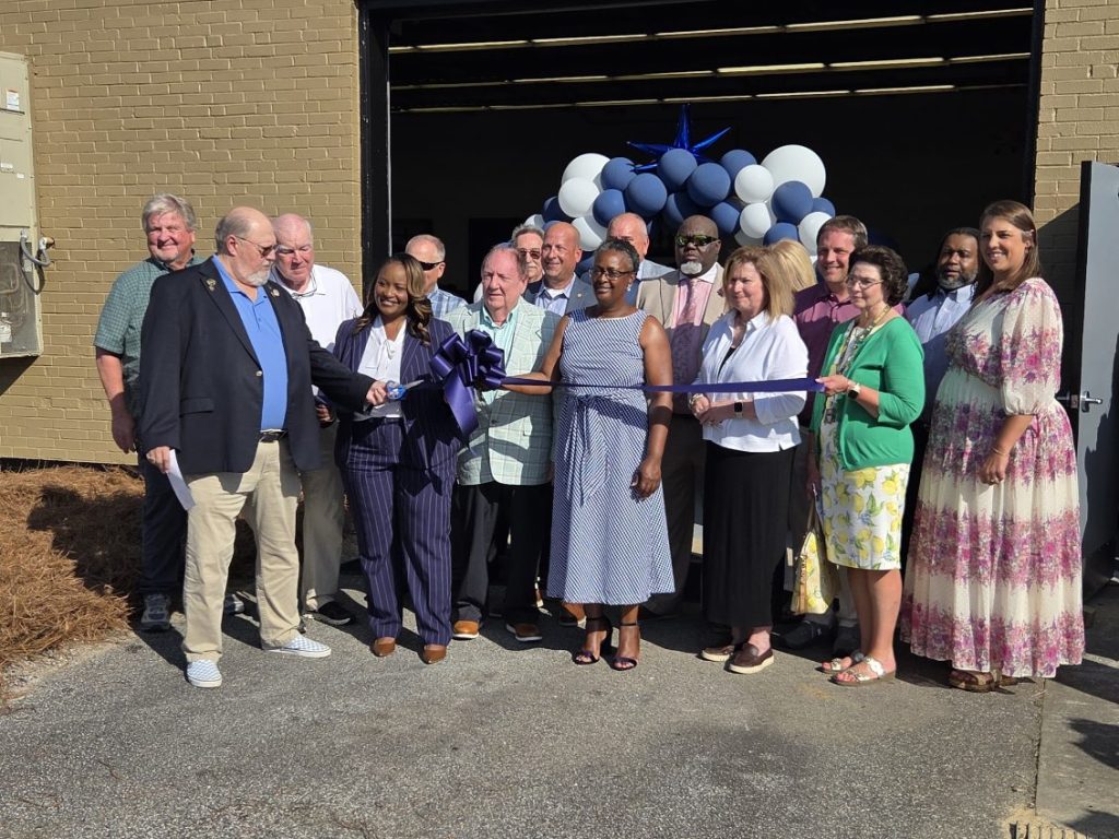 A group of people participate in a ribbon-cutting ceremony in front of a building entrance decorated with blue and white balloons.