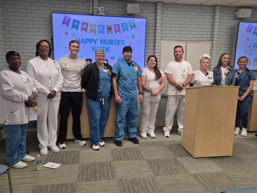 A group of nurses and staff standing together in a room, celebrating Nurses Week, with a "Thank You Happy Nurses Week" banner displayed on screens behind them.