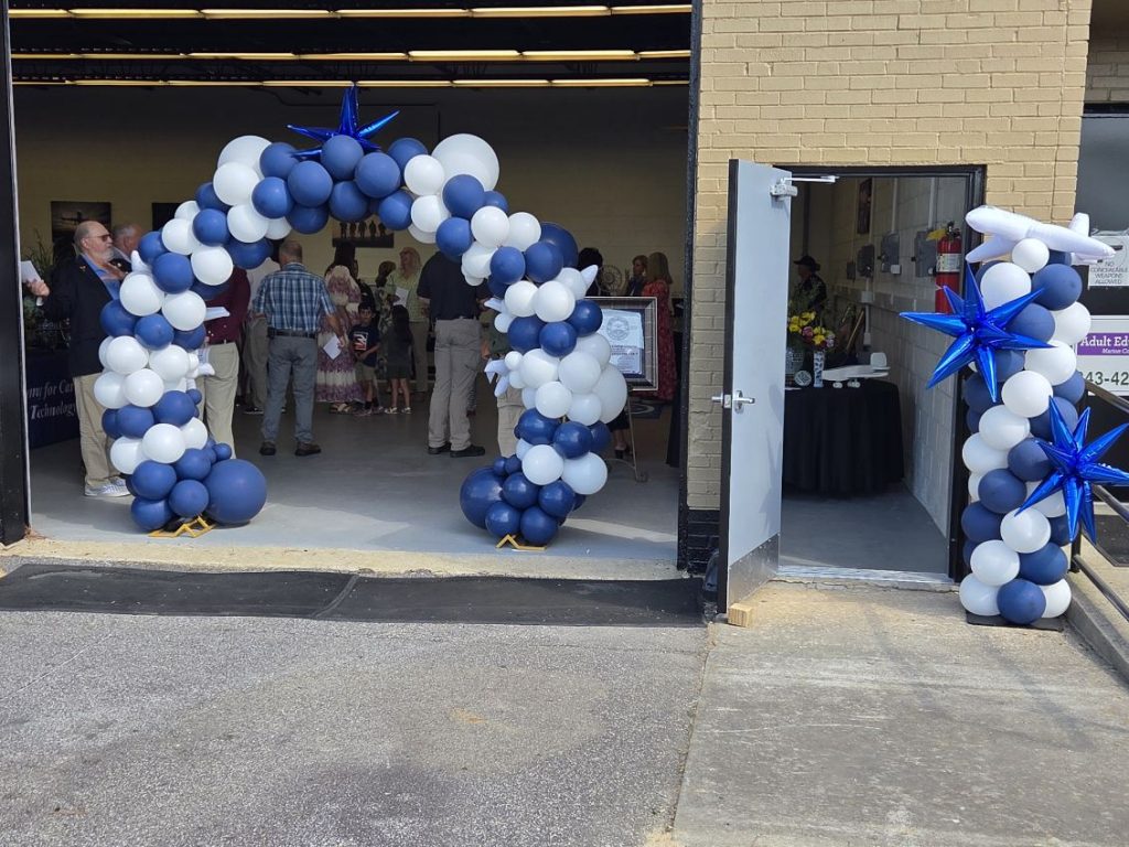 People gather inside a building decorated with blue and white balloon arches and star balloons, with tables, flowers, and signs visible near the entrance.