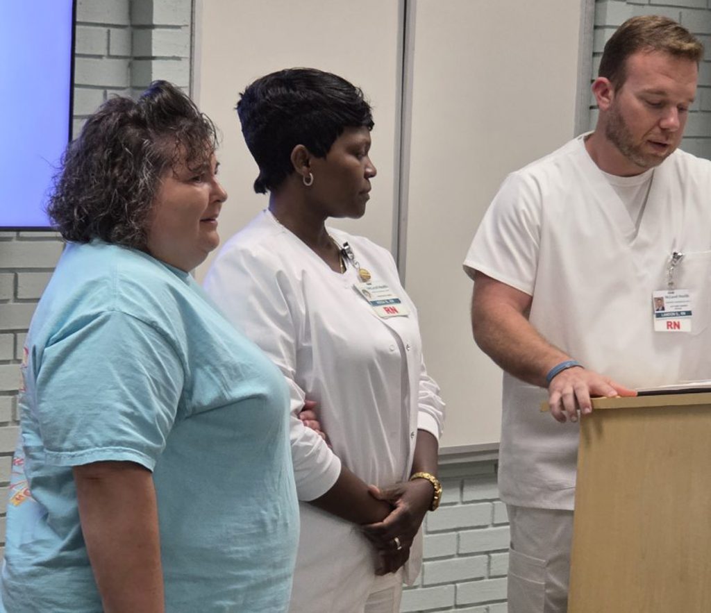 Three nurses stand together indoors, two in white uniforms with RN badges showing, while one reads at a wooden podium.
