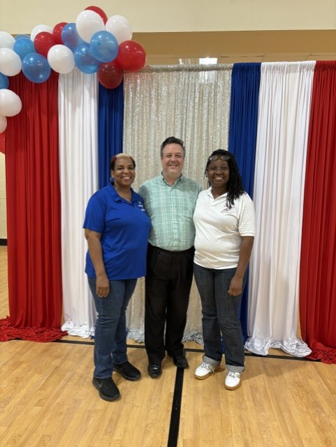 Three people stand smiling in front of red, white, and blue draped curtains and balloons in a gymnasium.