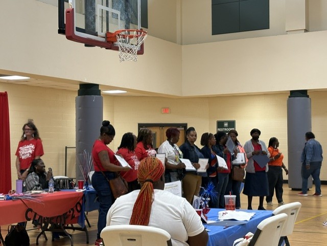 A group of people stand in a gymnasium holding certificates, while others sit at tables covered with tablecloths and refreshments.