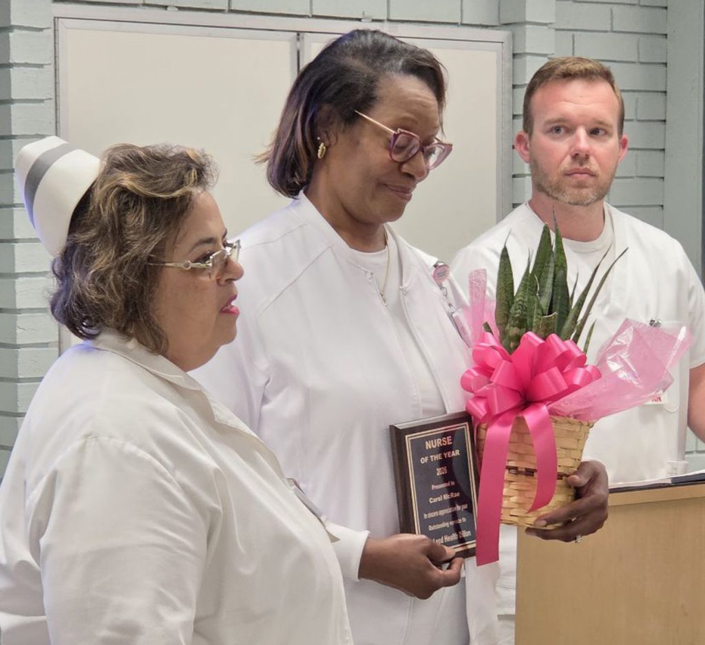 Three nurses in white uniforms, one holding a "Nurse of the Year" plaque and a plant with a pink ribbon, standing together indoors.