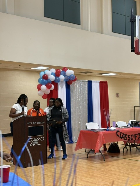 Three people stand at a podium in a decorated gym, with red, white, and blue balloons and banners, and a table with information materials nearby.