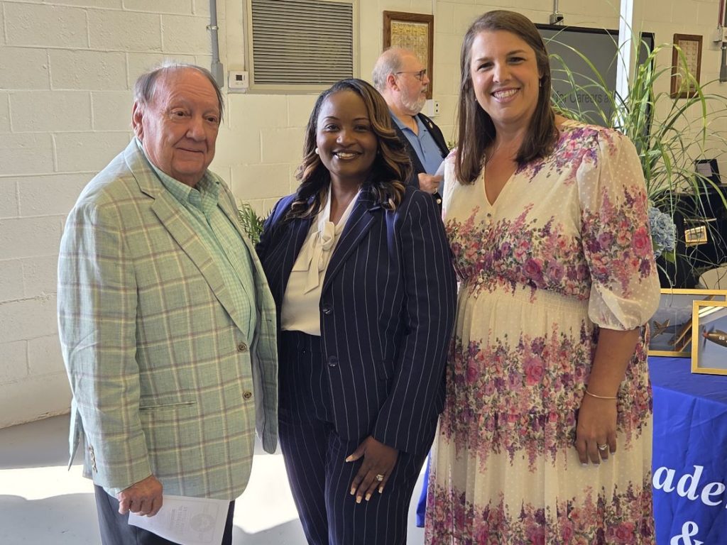Three people stand indoors, smiling at the camera. The man wears a checkered suit, the women wear a pinstripe suit and a floral dress. Plants and decor are visible.