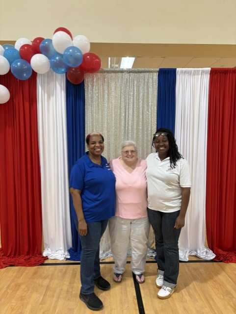 Three women stand together, smiling, in front of red, white, and blue curtains with matching balloons above, indoors.