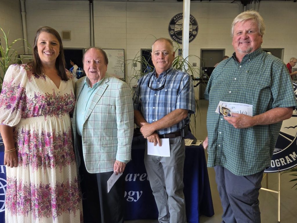 Four adults stand together indoors, smiling for a group photo in front of a table and sign, dressed in semi-formal and casual attire.