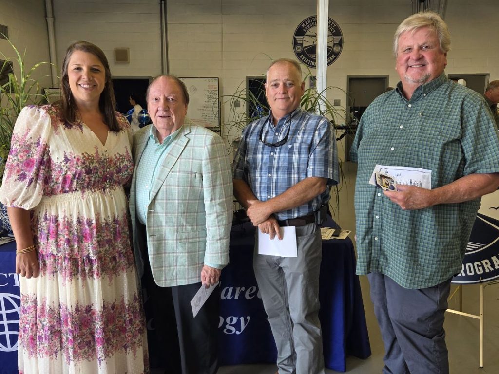 Four adults stand indoors, smiling at the camera, dressed in semi-formal attire. A table with informational pamphlets and plants appears in the background.