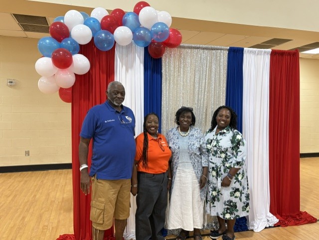 Four adults stand smiling in front of a red, white, and blue balloon and curtain backdrop indoors.