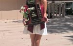Young woman holding bouquet and large softball championship trophy outside, wearing a white skirt and pink top, with columns and windows in the background.