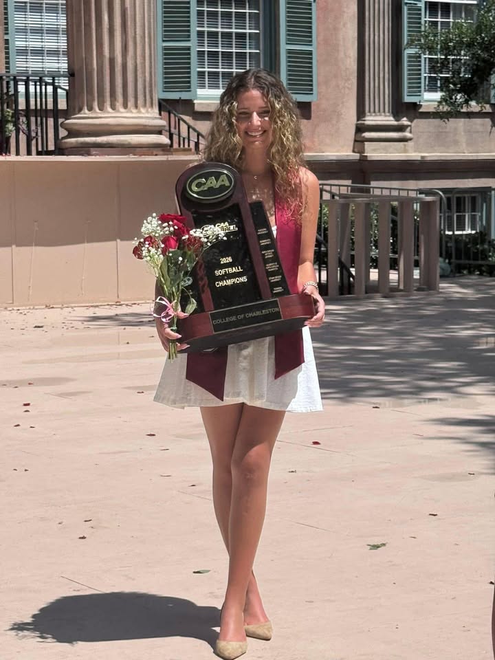 Young woman holding bouquet and large softball championship trophy outside, wearing a white skirt and pink top, with columns and windows in the background.