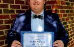 Person in a suit holding a certificate and smiling, standing in front of a brick wall.