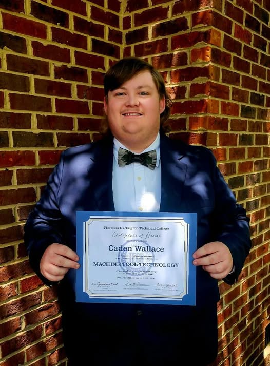 Person in a suit holding a certificate and smiling, standing in front of a brick wall.