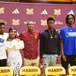 Five young adults stand together in a gym, smiling, with "Marion Swamp Foxes" and school logos on a maroon backdrop.