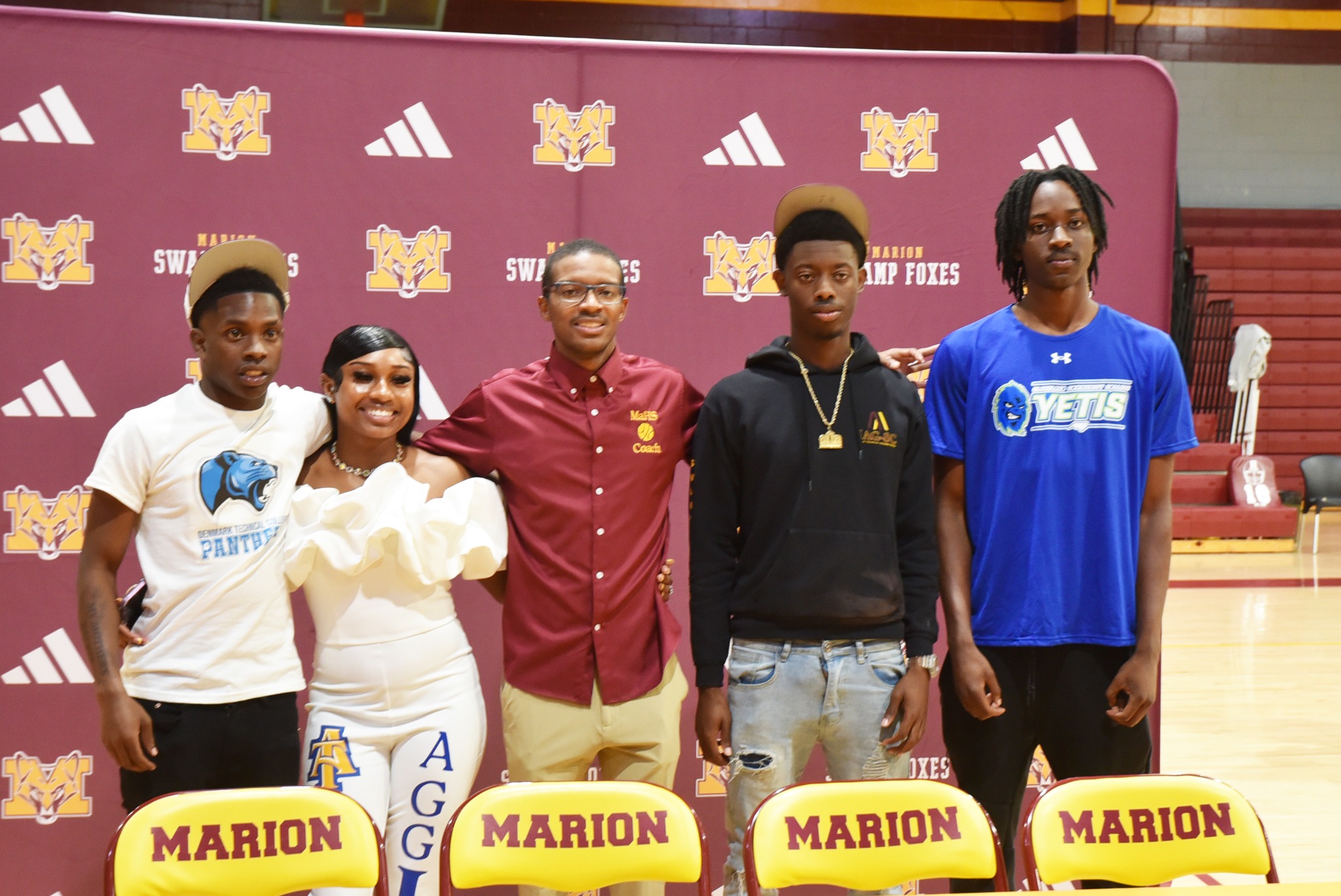 Five young adults stand together in a gym, smiling, with "Marion Swamp Foxes" and school logos on a maroon backdrop.