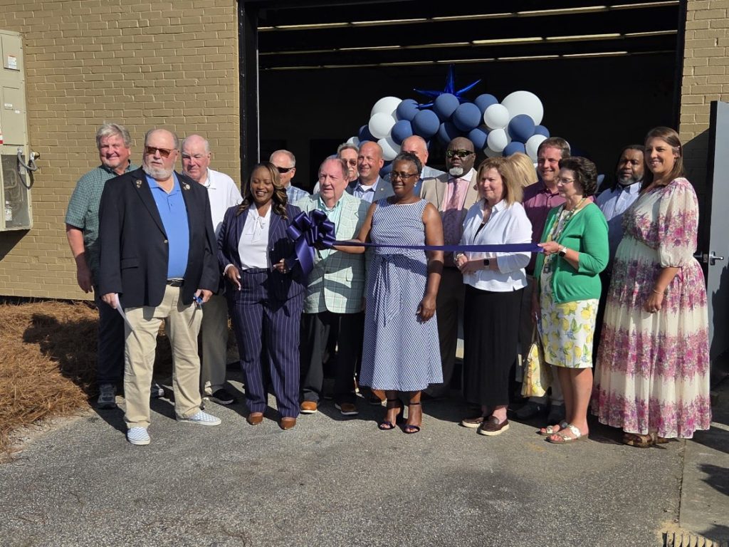 A group of people stands together outside a building, holding a ribbon for a ribbon-cutting ceremony, with balloons and decorations in the background.