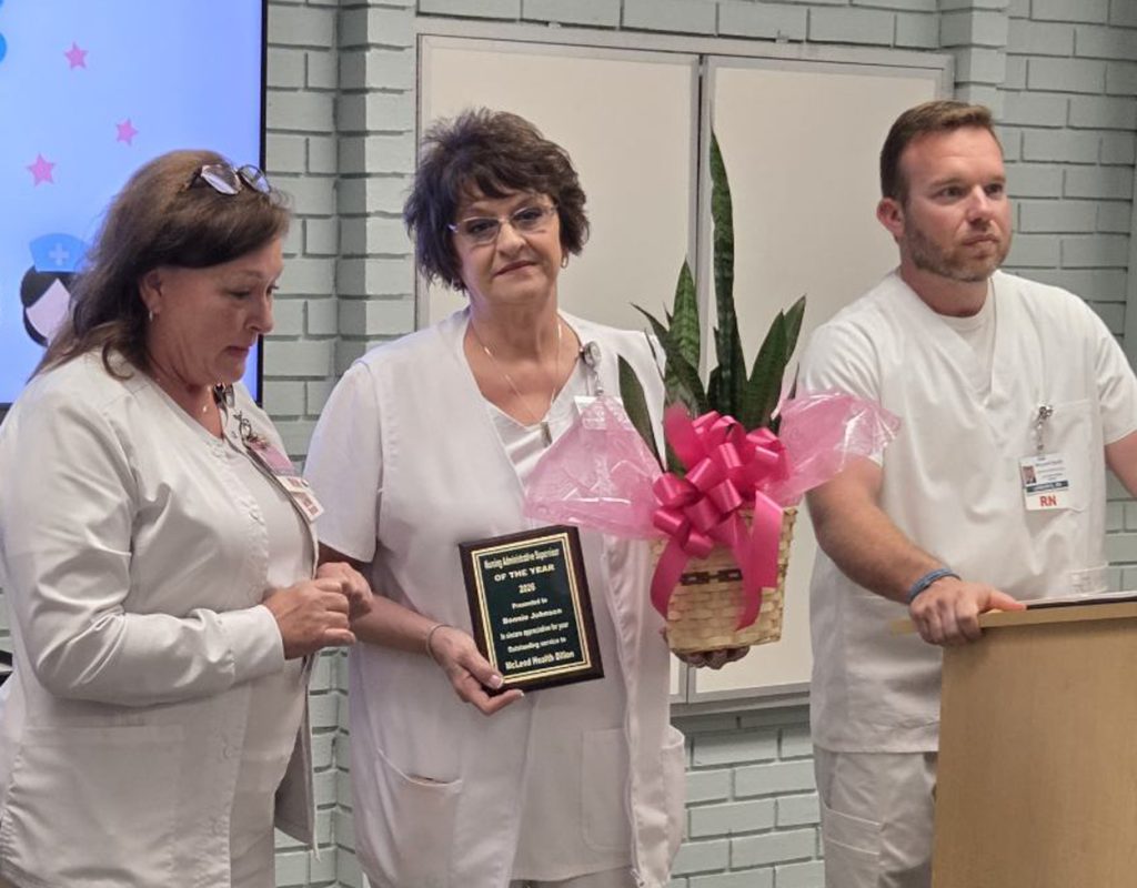 Three nurses in white uniforms stand together; one holds a plaque and a plant with a pink ribbon, appearing to receive an award in a hospital setting.