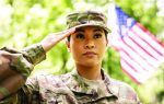 A woman in military uniform saluting outdoors, with an American flag in the background and trees surrounding the scene.