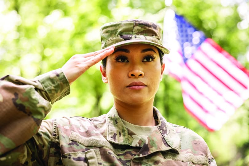 A woman in military uniform saluting outdoors, with an American flag in the background and trees surrounding the scene.